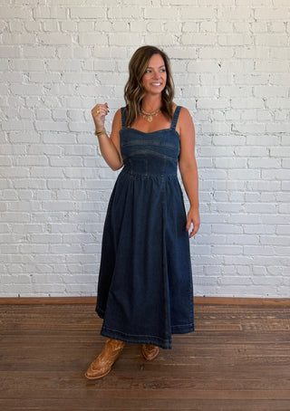 A woman wearing a dark blue denim midi dress with a smocked bodice and wide straps, standing on a wooden floor against a white brick wall.
