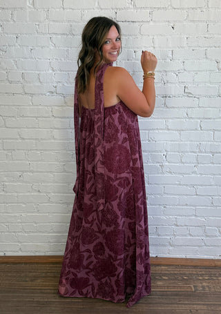 A woman wearing a sleeveless, floor-length burgundy maxi dress with a tonal floral pattern, standing in front of a white brick wall and wooden floor.