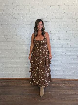 A woman wearing a brown tiered maxi dress with a beige floral pattern, standing in front of a white brick wall on a wooden floor.