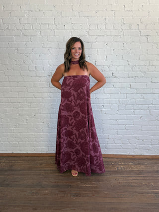 A woman stands against a white painted brick wall wearing a strapless, floor-length burgundy dress with a tonal floral pattern and a matching choker. The floor is dark wood.