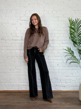 Woman wearing a brown sweater and black pants standing against a white brick wall with a plant to the right.