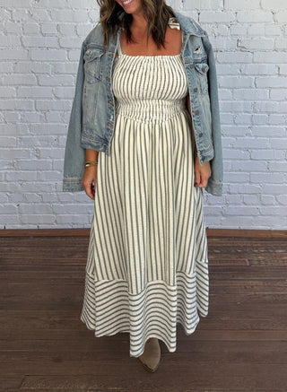 A woman modeling a cream and olive vertically striped maxi dress with a smocked bodice, styled with an open light-wash denim jacket, standing against a white brick wall and dark wood floor.