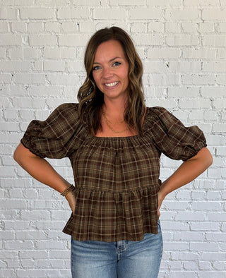 A woman modeling a brown, tan and taupe plaid puff-sleeve top with light-wash blue jeans, standing in front of a white brick wall and wooden floor.