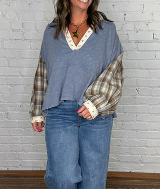A woman modeling a mineral washed denim top with plaid sleeves & floral hem paired with wide-leg blue jeans, standing against a white painted brick wall on a wooden floor.