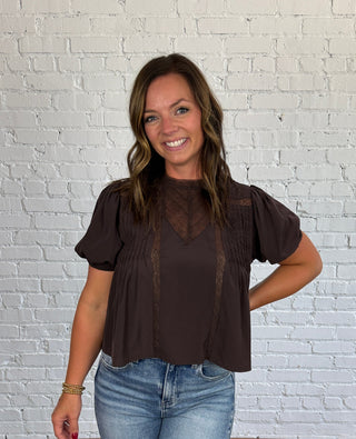 A woman modeling a dark brown short-sleeve blouse with sheer mesh puff sleeves and lace trim, paired with light blue jeans, standing against a white brick wall and wood floor.
