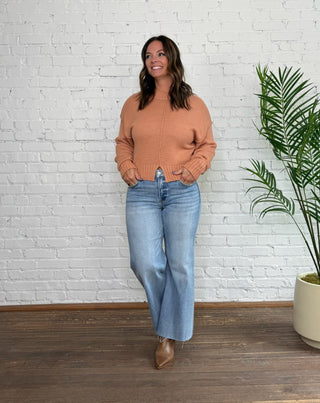 Woman wearing a brown sweater and blue jeans standing against a white brick wall with a plant to her right.