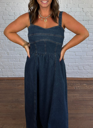 A woman modeling a dark blue denim jumpsuit in front of a white brick wall and wooden floor.