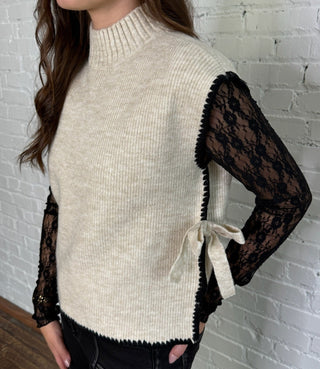 Woman wearing a cream sweater with black lace sleeves against a white brick wall.
