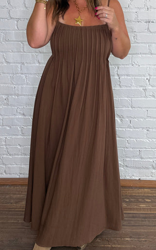 A woman modeling a brown sleeveless midi dress with a square neckline and thin straps, standing against a white brick wall on a wooden floor.