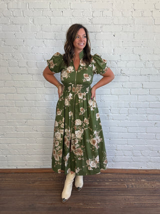 A woman modeling a green floral midi dress with puff sleeves and a front zipper, standing against a white brick wall on a wooden floor.