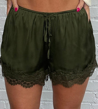 A woman stands against a white brick wall wearing olive green shorts, and brown cowboy boots.