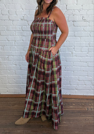 A woman modeling a thin strap maxi dress with pockets and a plaid pattern in burgundy, green, and blue, standing against a white brick wall on a wooden floor.