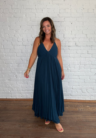 A woman modeling a deep V-neck, floor-length pleated maxi dress in dark teal blue, standing against a white painted brick wall on a wooden floor.