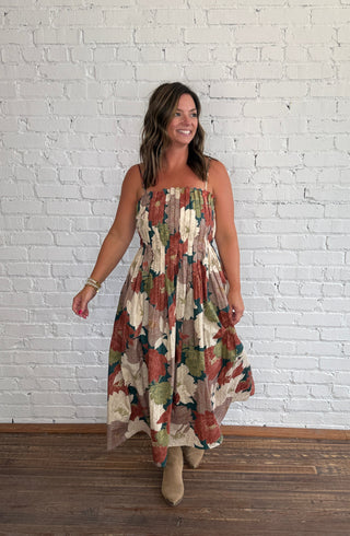 A woman wearing a spaghetti strap dress with a multicolor floral pattern in earthy tones, standing on a dark wood floor against a white brick wall.