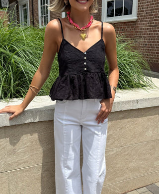 Woman in black top and white pants standing on a sidewalk with a brick building and greenery in the background.