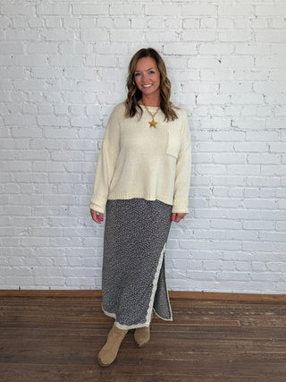 A woman posing in a cream knit sweater and a brown and blue floral midi skirt with cream lace trim, standing against a white brick wall and wooden floor.
