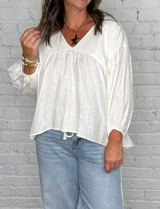A woman modeling a white long-sleeve v-neck blouse and light-wash blue jeans, standing against a white painted brick wall on a dark wood floor.