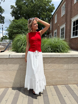 Woman in a red top and white skirt standing outdoors near a brick building.