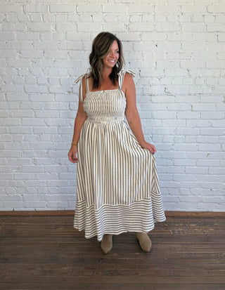 A woman modeling a sleeveless, square-neck maxi dress with vertical stripes in cream and brown, standing against a white brick wall on a dark wood floor.