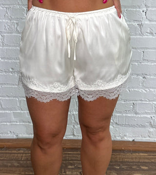 A woman stands in front of a white brick wall wearing white shorts with a lace hem, and beige ankle boots.