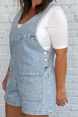 A woman stands in front of a white brick wall wearing a short, light blue denim overall romper with a horseshoe embroidered on each pocket, a white t-shirt underneath, and brown cowboy boots.