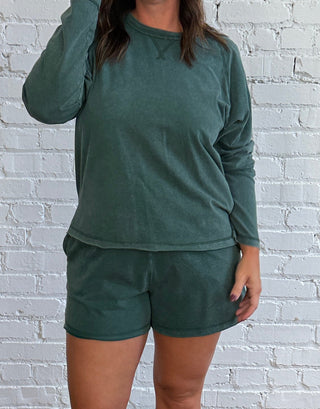 A woman modeling a matching dark green lounge set consisting of a long-sleeve shirt and shorts.