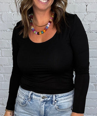 A woman stands in front of a white brick wall wearing a fitted black long-sleeve top, light-wash blue jeans, and brown shoes, with a colorful beaded necklace.