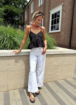 Woman in black top and white pants standing on a sidewalk with a building and greenery in the background