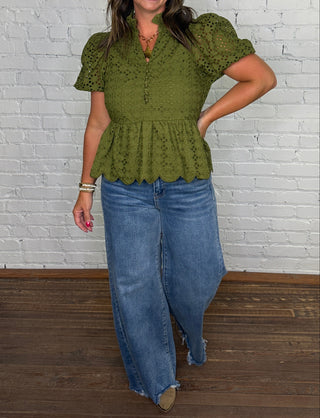 A woman modeling an olive green eyelet peplum top with puff sleeves and blue wide-leg jeans, standing against a white painted brick wall on a dark wood floor.