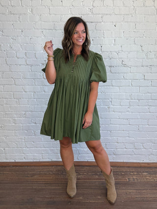 A woman modeling an olive green pleated mini dress with puff sleeves and tan ankle boots, standing against a white brick wall and dark wood floor.