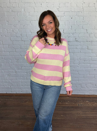 A woman wearing a pink and yellow striped knit sweater, blue jeans, and brown shoes, posing against a white brick wall and dark wood floor.