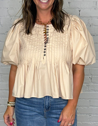 A woman modeling a cream-colored short-sleeve blouse with puff sleeves and a button-up front, paired with wide-leg blue jeans, standing against a white brick wall and wooden floor.