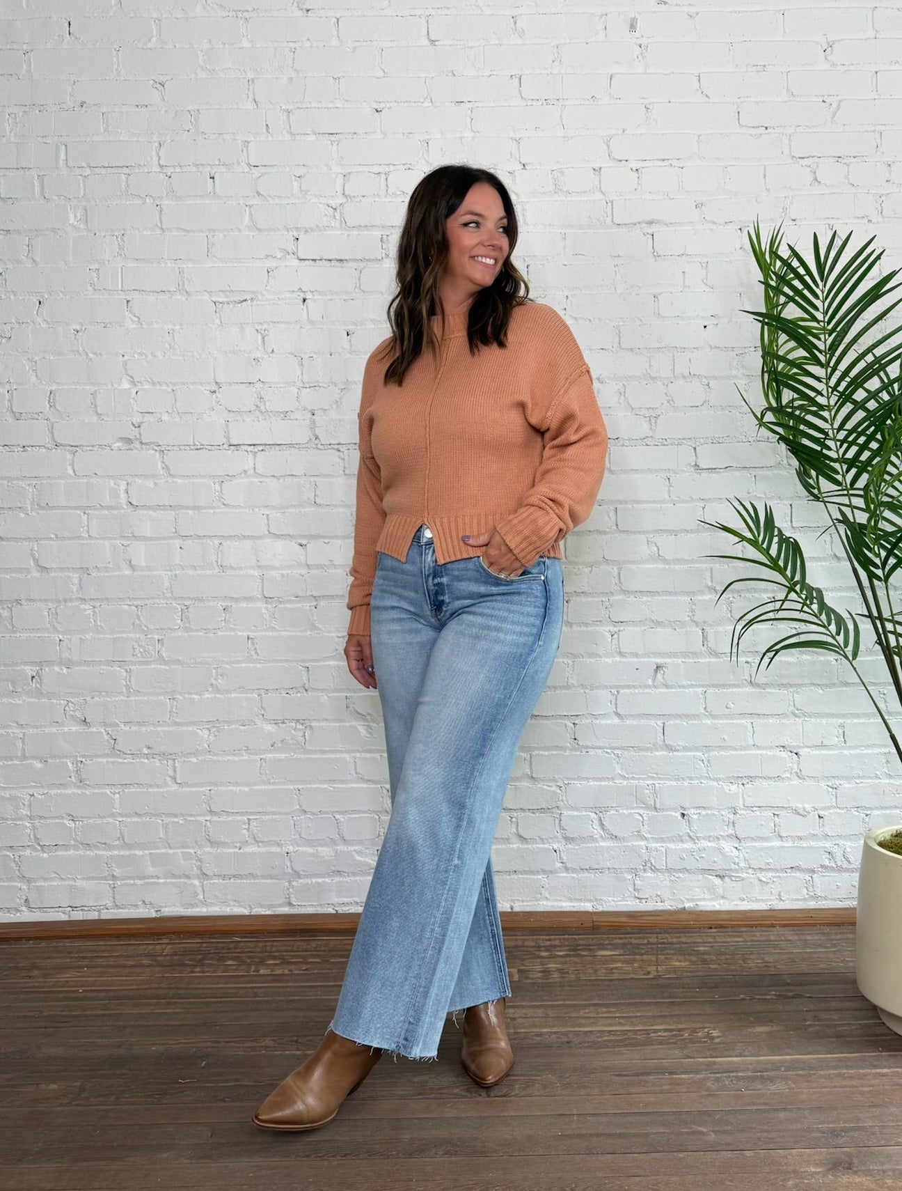 Woman wearing a brown sweater and blue jeans standing against a white brick wall with a plant to her right.