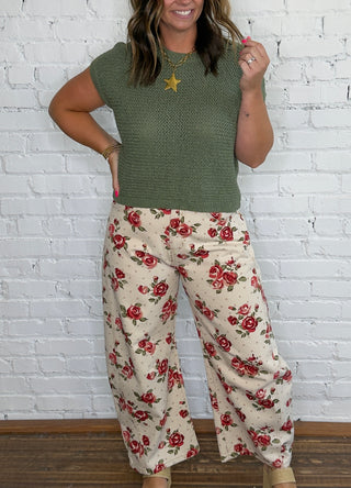A woman wearing a textured olive green short-sleeve knit top and cream wide-leg pants with a red rose floral print.