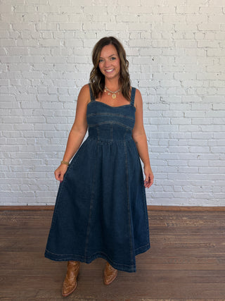 A woman modeling a dark blue denim midi dress with a corset-style bodice and A-line skirt, standing against a white painted brick wall on a wooden floor.