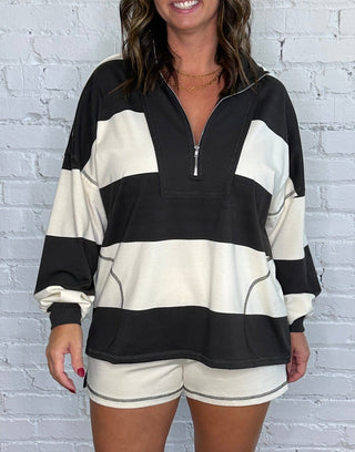 A woman modeling a charcoal and cream striped half-zip sweatshirt and matching shorts set, standing on a wooden floor against a white brick wall.