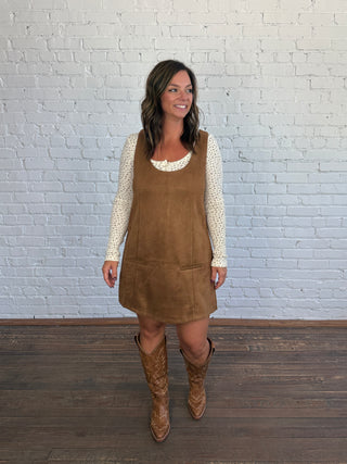 A woman modeling a brown dress over a cream, floral, long-sleeve top, paired with brown cowboy boots, standing against a white brick wall.