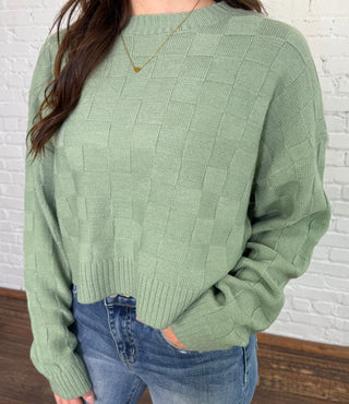 Person wearing a green sweater and blue jeans sitting on a wooden floor.