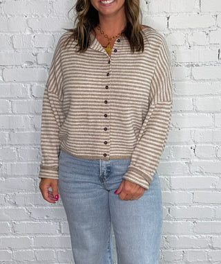 A woman stands against a white brick wall on a dark wood floor, wearing a beige and brown horizontally striped long-sleeve button-up shirt, light-wash wide-leg jeans, and tan ankle boots.