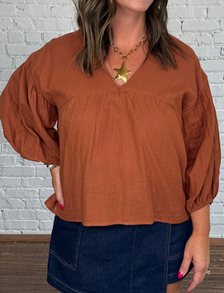 A woman modeling a rust-orange long-sleeve blouse with a dark denim mini skort, standing against a white brick wall.