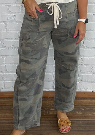 A woman stands against a white brick wall wearing a cream ribbed sleeveless top with gold buttons and gray camouflage wide-leg pants, paired with tan woven slide sandals.