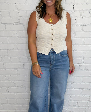 A woman wearing a cream sleeveless button-front top and wide-leg blue jeans, with a gold pendant necklace and sandals.