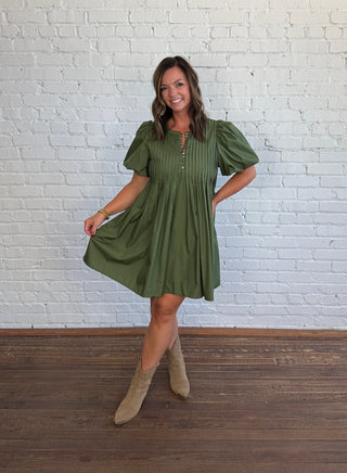 A woman modeling an olive green puff-sleeve mini dress with a gathered waist and front button placket, paired with beige ankle boots, standing against a white brick wall and dark wood floor.
