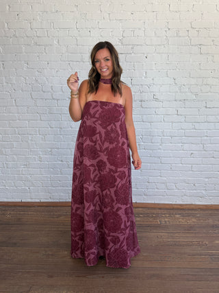 A woman modeling a strapless maxi dress and matching scarf with a floral pattern in deep burgundy and plum, standing against a white brick wall and wooden floor.