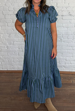 A woman wearing a blue and green vertically striped midi dress with puff sleeves & pockets paired with beige ankle boots, standing against a white brick wall on a wooden floor.