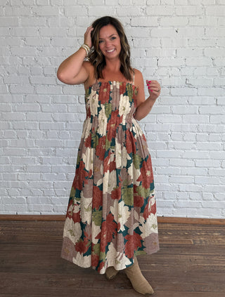 A woman wearing a spaghetti strap dress with a multicolor floral pattern in earthy tones, standing on a dark wood floor against a white brick wall.
