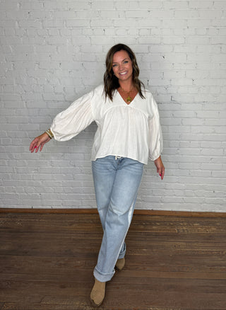 A woman posing in a white long-sleeve blouse with balloon sleeve, light-wash blue jeans, and tan boots, standing against a white brick wall and wooden floor.
