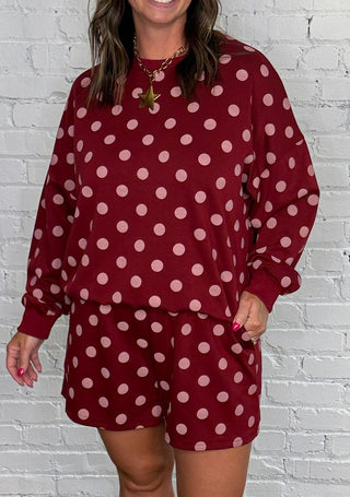 A woman modeling a matching burgundy and pink polka dot two-piece set standing against a white brick wall and wooden floor.
