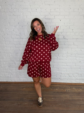 A woman modeling a matching burgundy and pink polka dot two-piece set standing against a white brick wall and wooden floor.