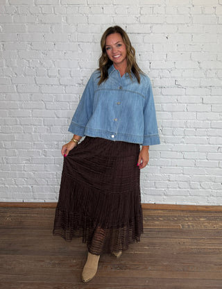 A woman stands in front of a white brick wall wearing a light blue cropped denim jacket and a dark brown lace tiered midi skirt, paired with taupe ankle boots.